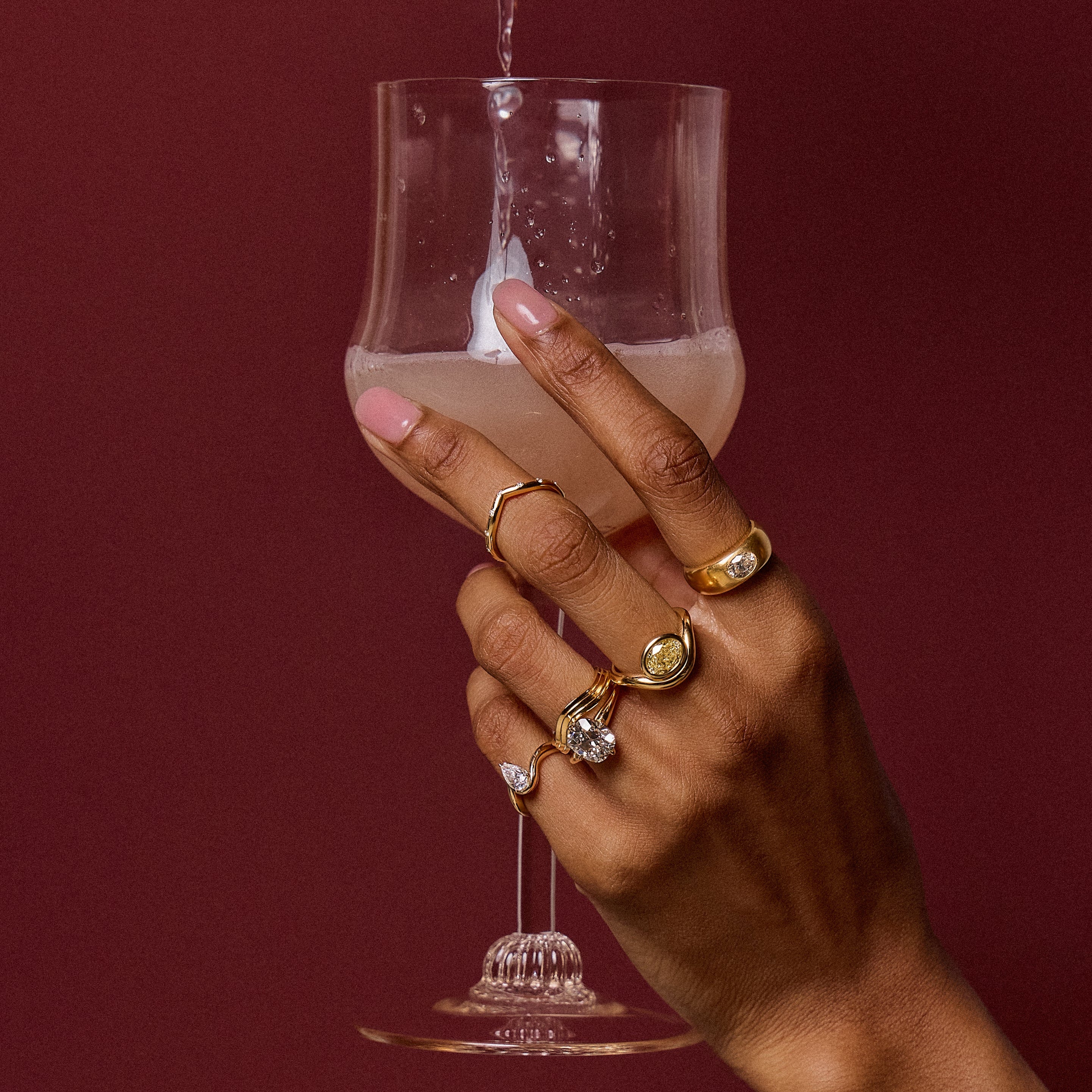 Hand model holding a pink wine glass in front of a burgundy background wearing BB Talia White Diamond in 14k yellow gold on left pinky, MAE 3.53 RING in 18k gold and KYMA Band in 18k yellow gold on ring finger, IRIS band in 18k gold and DEEPA 2.04 Ring in 18k yellow gold on middle finger.